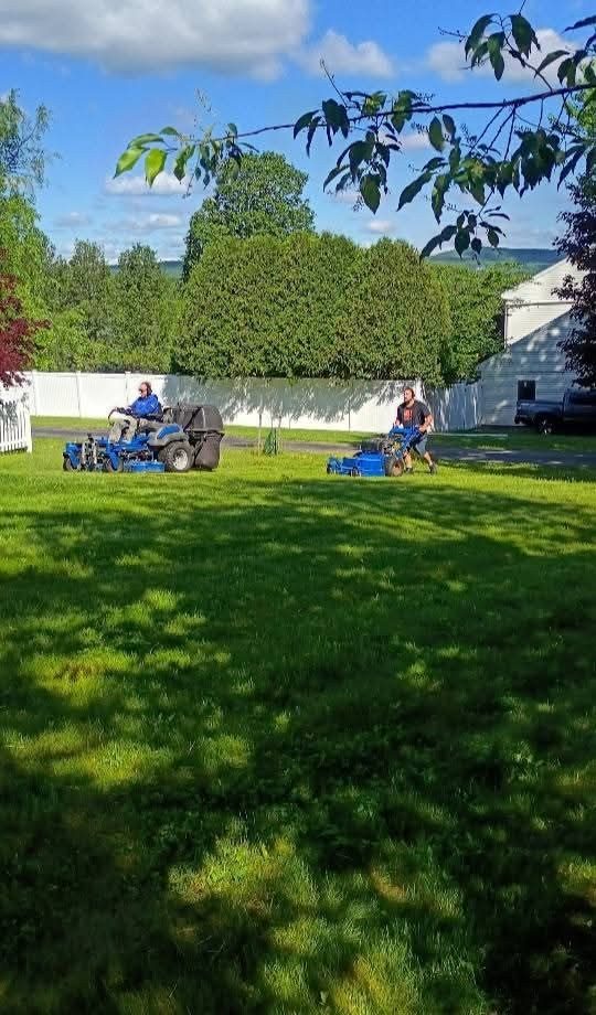 Two blue mowers working a yard by a white fence